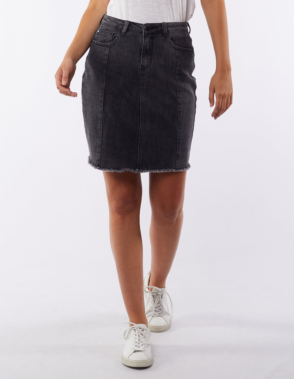A person is wearing a black Foxwood Kiama Skirt with a frayed hemline, paired with a white T-shirt and white sneakers. The person is in mid-step, with their left arm slightly bent and their right arm hanging by their side, capturing an effortlessly casual vibe against the plain white background.