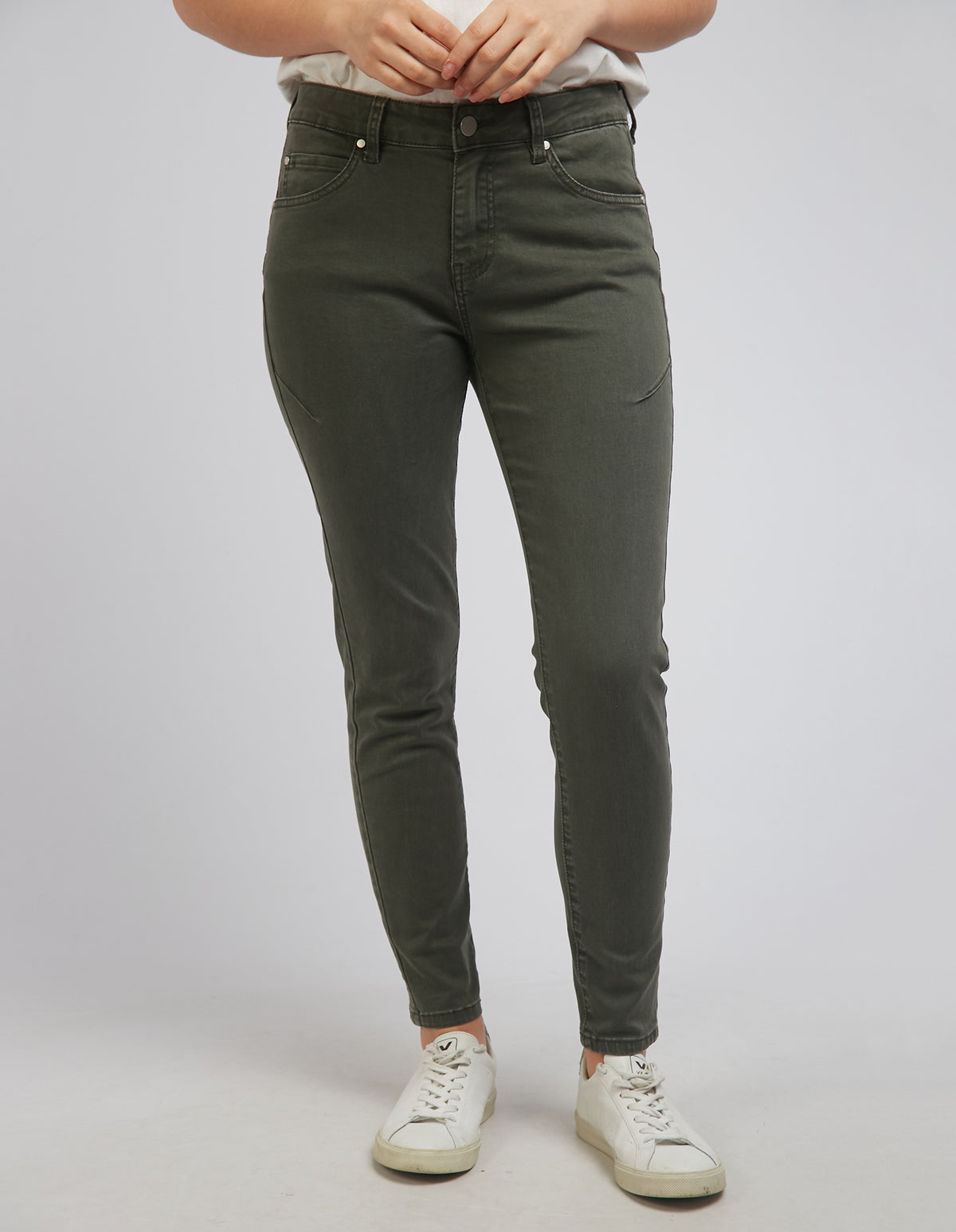 A person wears the Foxwood City Jean, high-waisted in olive-green cotton with a skinny leg fit, paired with white sneakers and a light top. Their hands rest by their sides against a plain background.