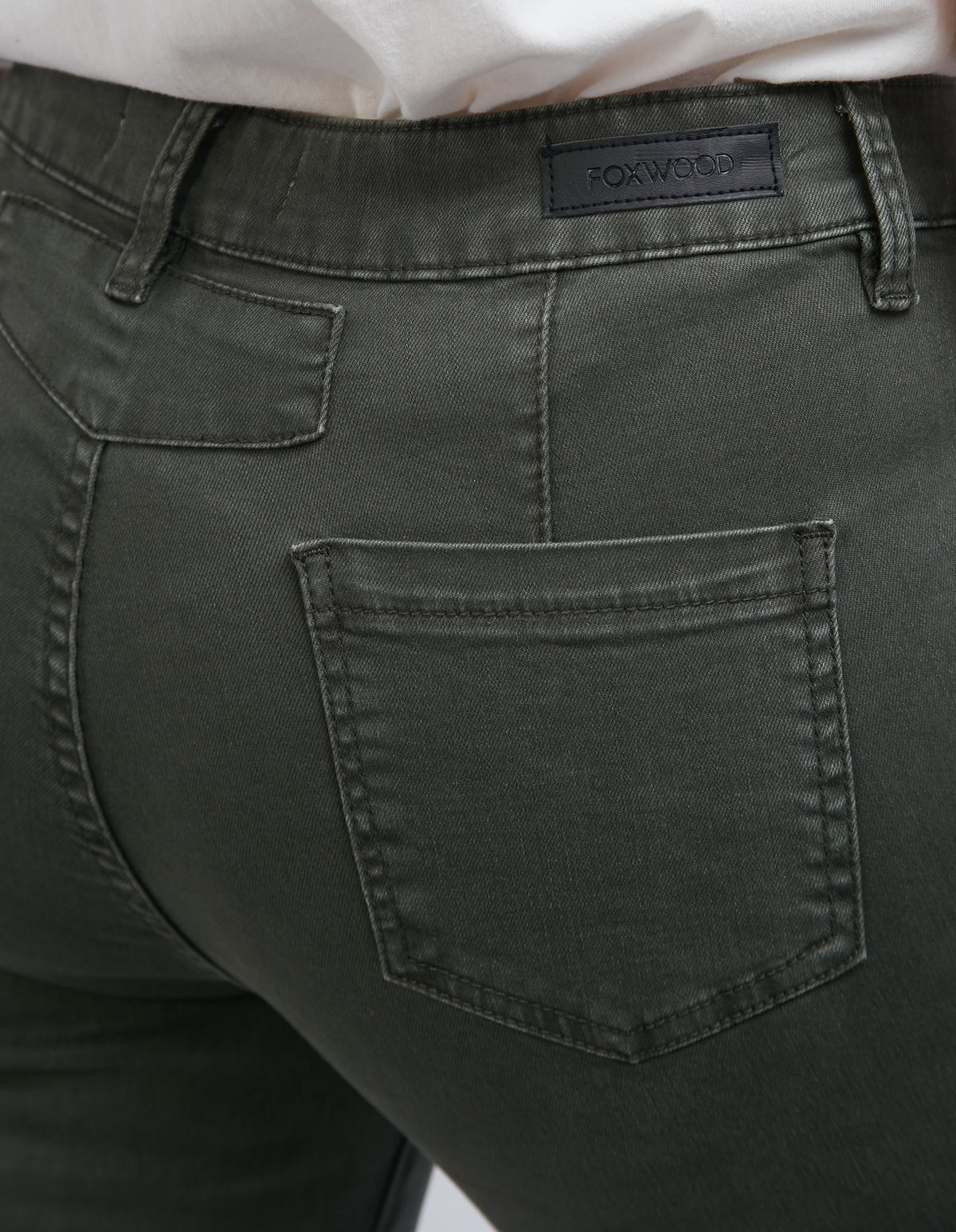 Close-up of someone in high waist dark green Foxwood City Jean, featuring visible back pockets and belt loops. A black "FOXWOOD" label is on the waistband. The skinny leg fit goes well with their white top.