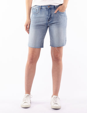 A person wearing the Foxwood AJA Bermuda Shorts in light blue denim with super soft, distressed detailing and a white t-shirt. Their left hand is in their pocket, and they are standing against a plain white background. The person is also wearing white sneakers.