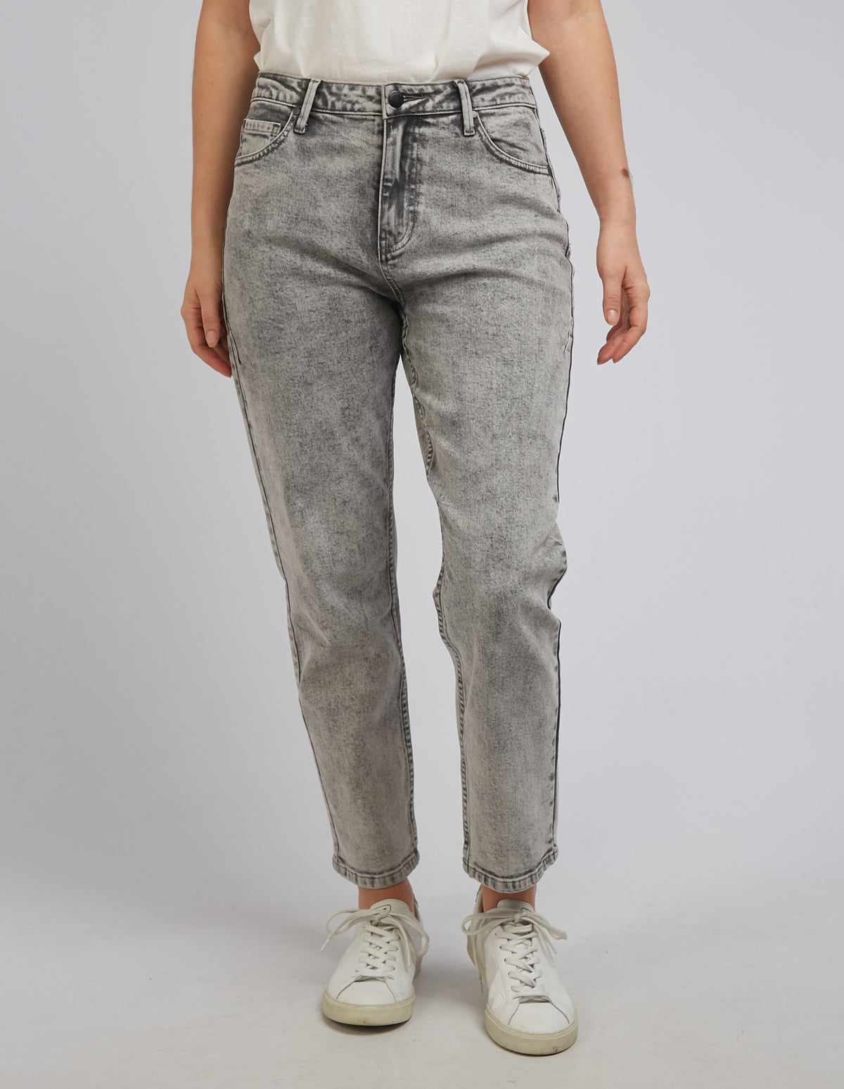 Dressed in white sneakers and a white t-shirt, an individual is posing against a plain gray background in the comfortable Foxwood Barkly Straight Leg Jeans, featuring a subtle patch. The stylish jeans enhance their relaxed stance with arms at their sides.