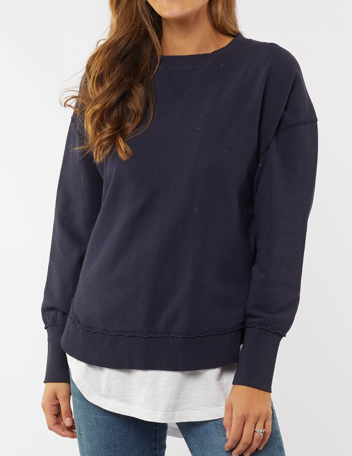 A person dons the Foxwood Delilah Crew, a navy blue sweatshirt with raw edge detailing, over a white shirt and blue jeans. With long wavy hair obscuring their face, they stand mysteriously against a plain light background.