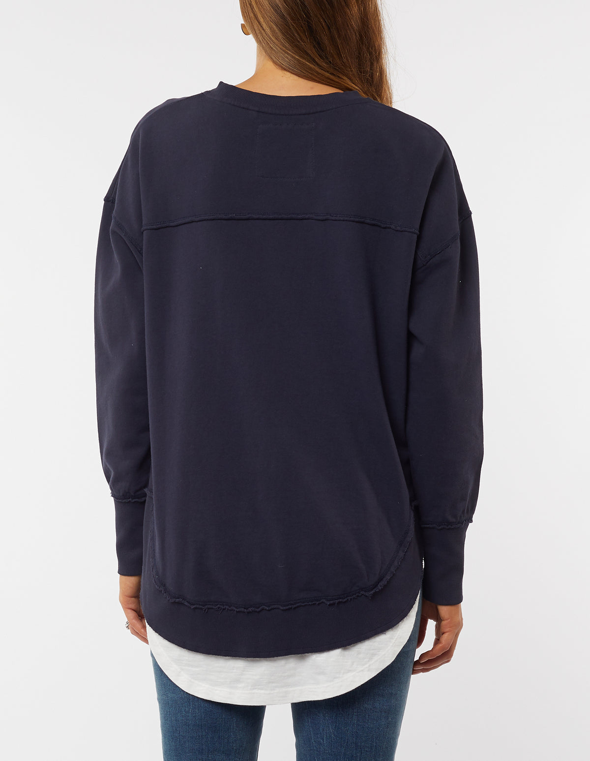 A person with long hair wears a navy blue Foxwood Delilah Crew, featuring raw edge detailing, over a white shirt. The 100% cotton oversized jumper pairs perfectly with blue jeans as they turn away, highlighting the sweatshirt's back.