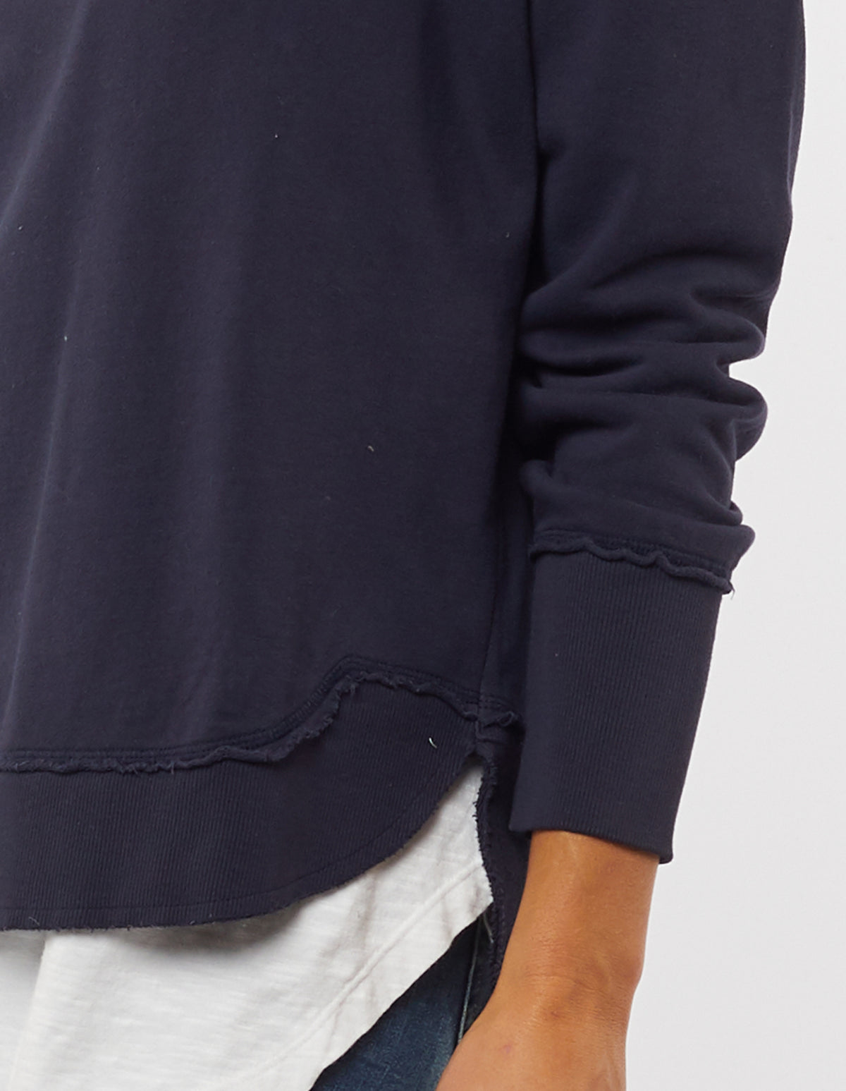 A close-up of a person wearing the Foxwood Delilah Crew, a navy blue cotton jumper with a scalloped hem and ribbed cuffs, layered over a white shirt. The person's hand rests at their side.