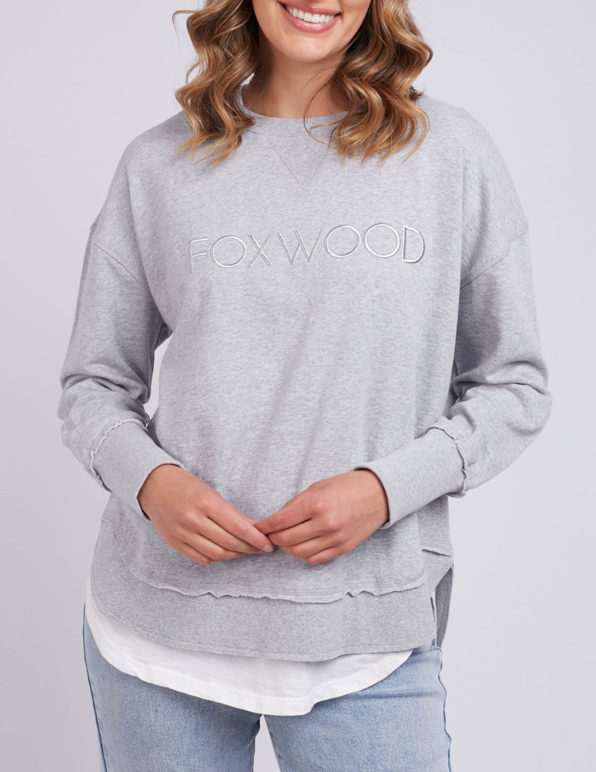 A person smiles while wearing the Foxwood Simplified Crew in light gray, paired with light blue jeans. Their wavy, shoulder-length hair stands out against a plain gray background.