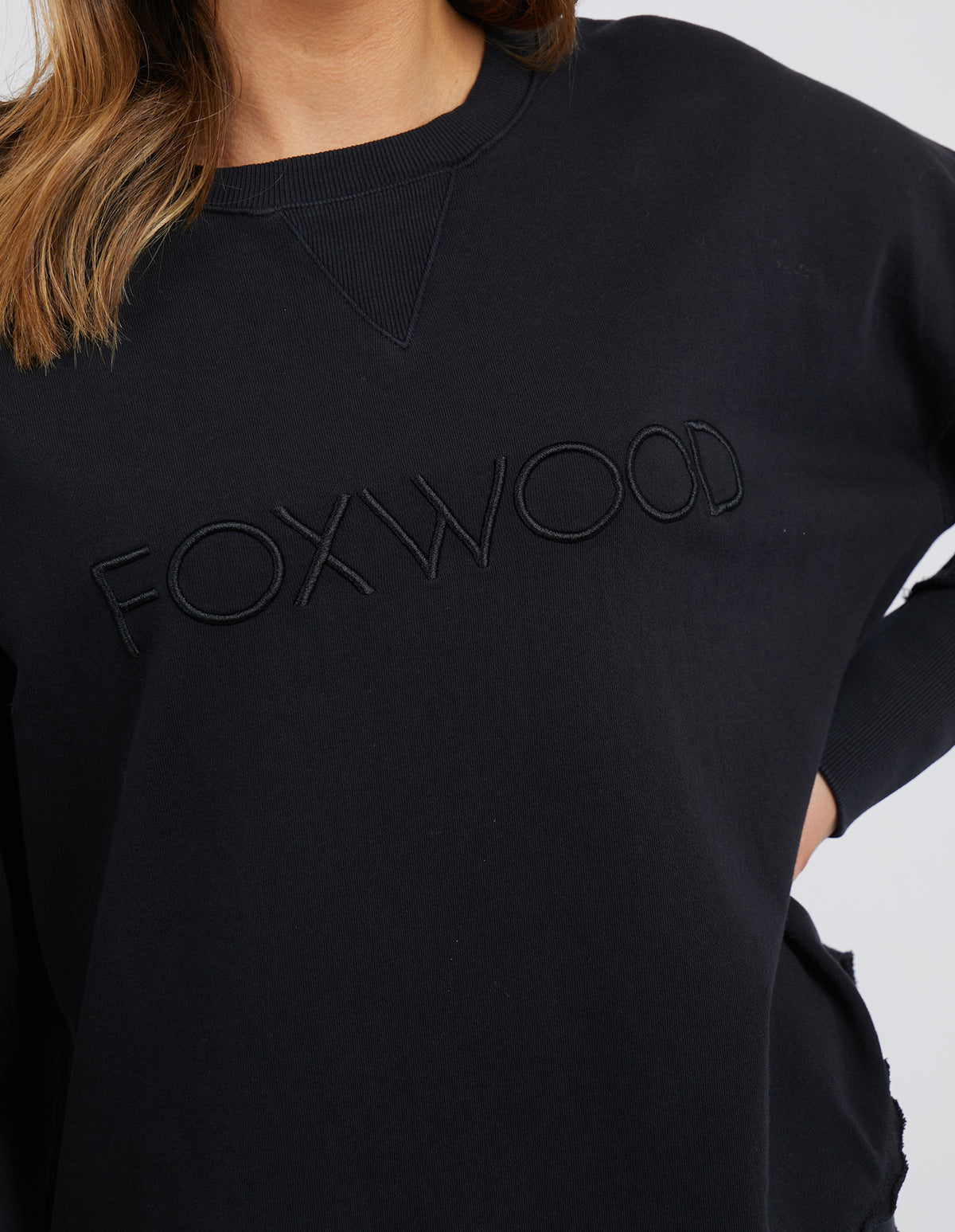 A person is seen wearing a black Foxwood Simplified Crew sweatshirt with "FOXWOOD" embroidered on the front, highlighting its relaxed fit. Only the upper torso and part of the left arm are visible against a plain white background, emphasizing its 100% cotton fabric.