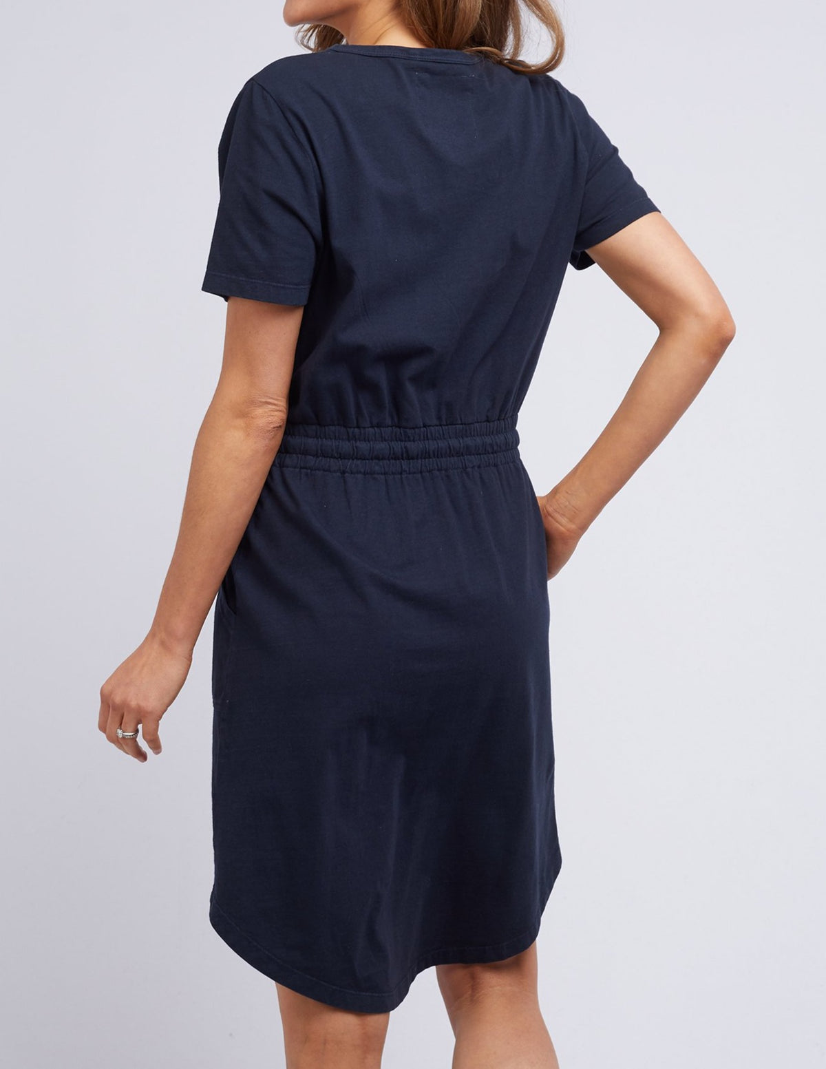A person is seen from the back wearing a navy Foxwood Ingrid dress, featuring short sleeves and an elastic waist. They stand with one arm slightly bent against a plain light gray background.