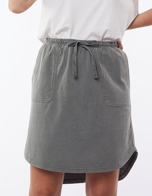 A person wearing a white top is shown from the waist down. They are wearing the Foxwood Palm Skirt, a comfortable gray knee-length skirt made of 100% Cotton Baby Cord. The skirt features an elastic waistband with a drawstring, two front pockets, and a slightly curved hemline at the sides.