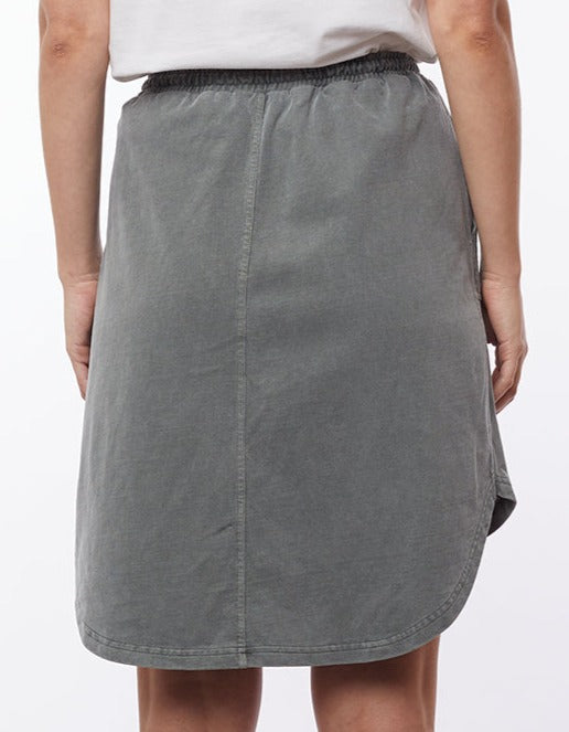 A person is standing with their back to the camera, wearing the Foxwood Palm Skirt—a gray, knee-length piece featuring an elastic waist and rounded hem. Crafted from 100% Cotton Baby Cord, this comfortable skirt pairs effortlessly with a light-colored top that peeks out above the waistband. The background is white.