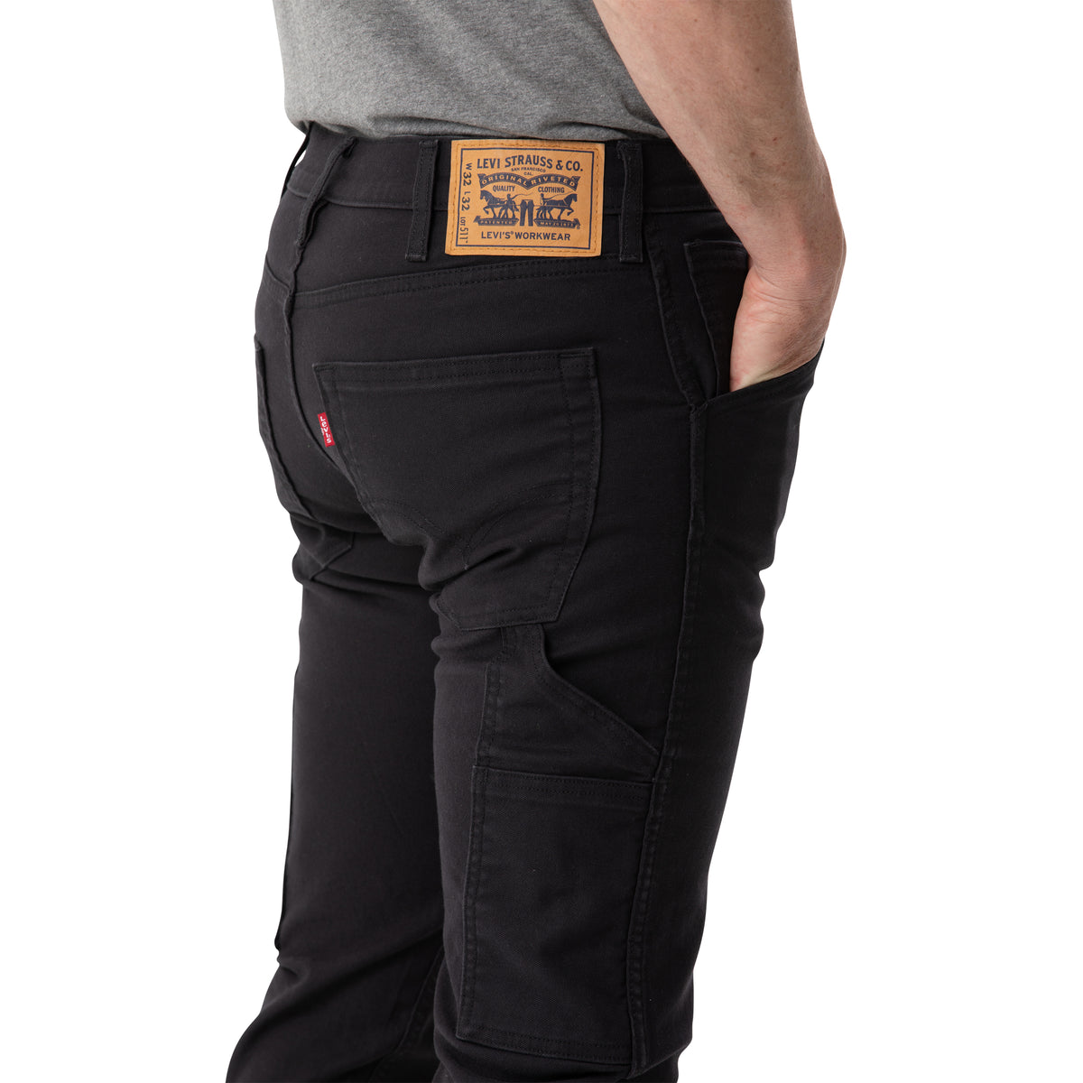 A person wearing black Levis Workwear 511 Utility Slim Jean with a visible brown Levi Strauss & Co. leather patch on the waistband, stands with their right hand in their pocket. They are pairing the stretch jeans with a grey shirt tucked into them. The image focuses on the person's lower back and legs.