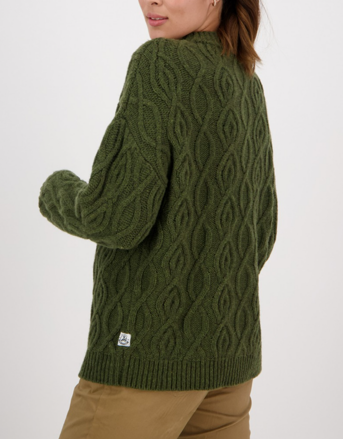 A person in khaki pants and an olive green Swanndri Pendeen Cable Neck Crew, featuring a chunky cable-knit texture and a small white label near the hem, stands turned slightly away, looking over their shoulder.