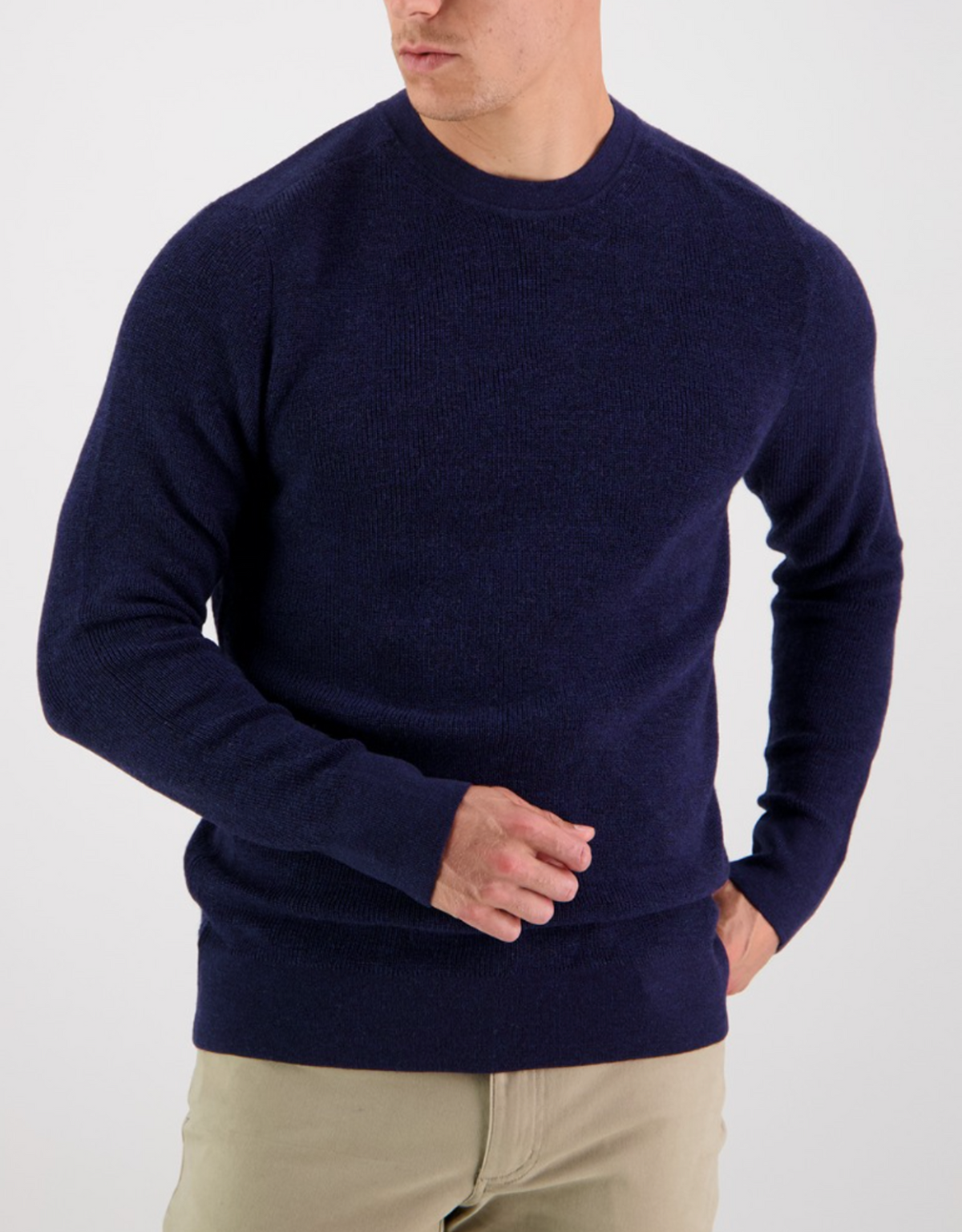 A person poses against a white background wearing the Swanndri Otahu River Crew Neck Merino Knit in navy blue and beige pants. They turn slightly to the side, elegantly adjusting the sweater with one hand.