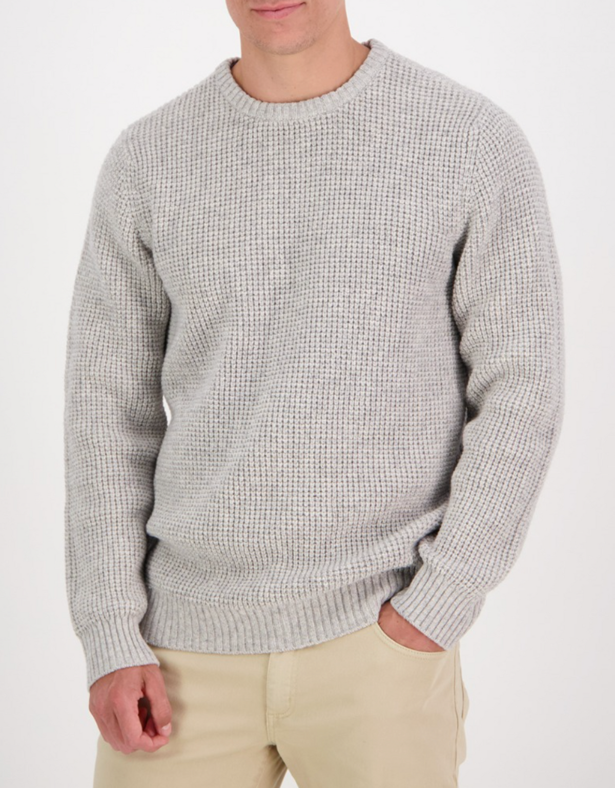 A man wears a light gray Swanndri Fistral Waffle Knit lambswool sweater, paired with beige pants against a plain white background. His hand rests casually in his pocket, with his head slightly cropped out of frame.