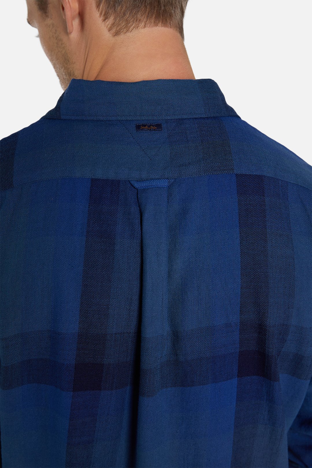 Close-up of the back of a man's head and shoulders, focusing on an Industrie The Hertford Shirt in a blue checkered lightweight fabric. The shirt features a box pleat, long sleeves, and a locker loop on the upper back. The man's head is turned slightly to the side, showcasing the Industrie logo and the detailed craftsmanship of the shirt.