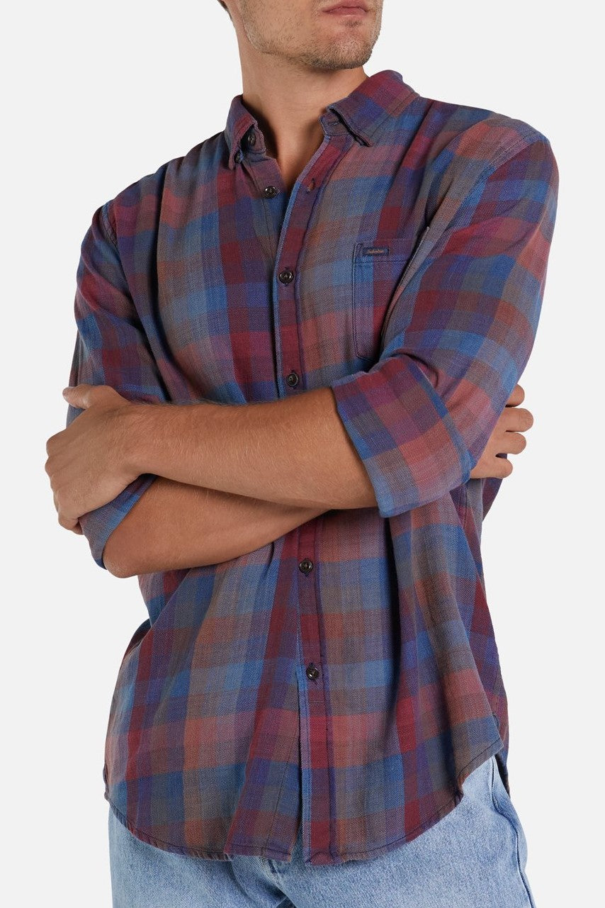 A person stands with arms crossed, wearing the Industrie The Harlow Shirt, a long-sleeved, button-up plaid shirt in shades of blue, maroon, and red. The lightweight shirt is slightly crinkled and untucked, paired with light blue jeans. The person is looking to the side.