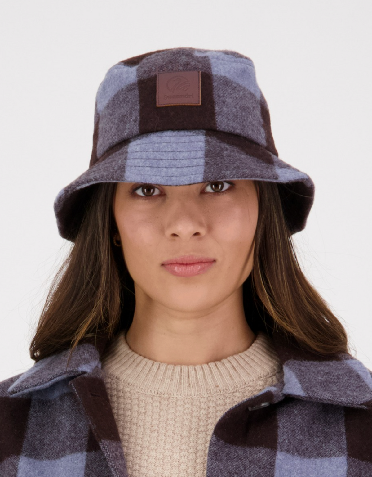 A person wears a Swanndri Kaimai Wool Bucket Hat in blue and brown check, paired with a matching jacket and beige merino sweater, looking at the camera. The hat has a leather patch logo on the front against a plain white background.