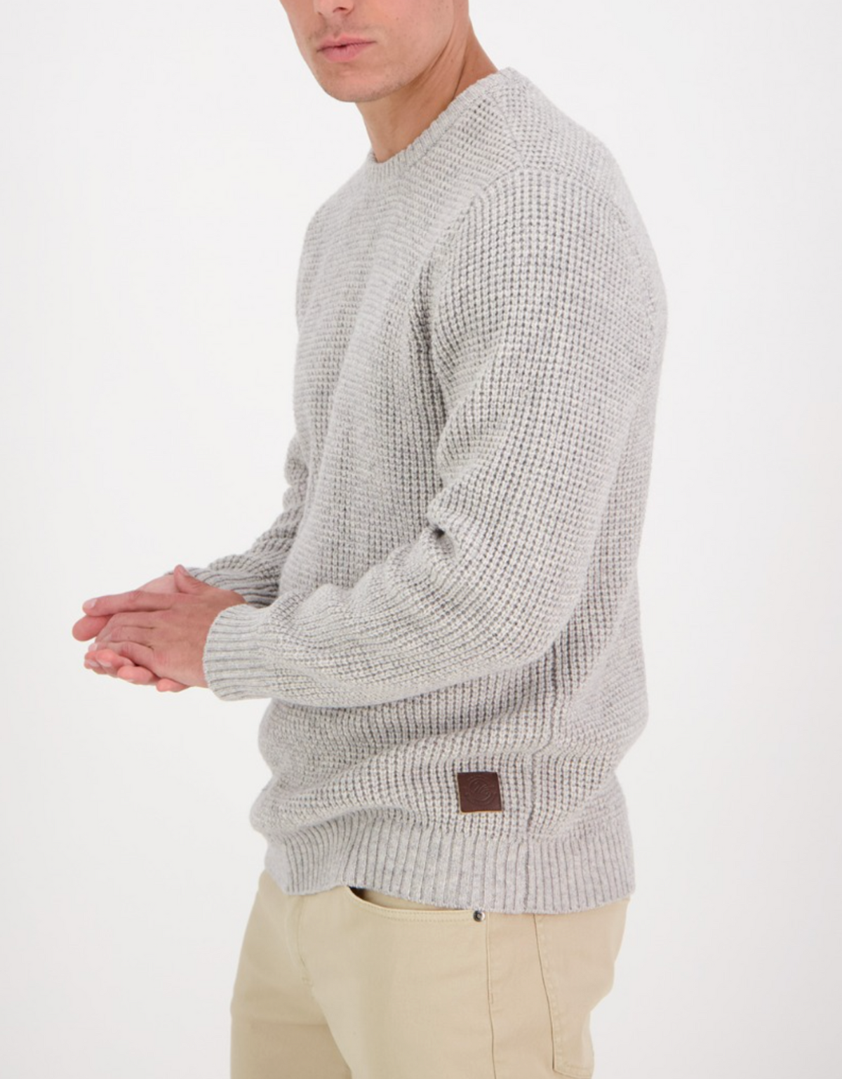 A person modeling the Swanndri Fistral Waffle Knit in light gray is posing against a white background, wearing it with beige pants and hands clasped. The sweater features a small leather patch near the hem, with their face partially obscured.