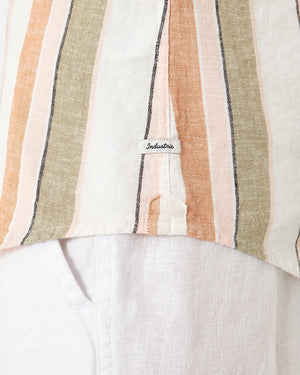 Close-up of a person's torso wearing a casual ensemble featuring Industrie The Orchard Linen Shirt and white pants. The long-sleeved shirt, created from 100% linen, showcases vertical stripes in shades of peach, beige, and olive. A small tag near the hem reads "Industrie." The white pants exhibit a visible side seam and pocket.