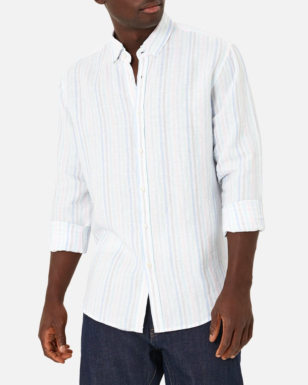 A person in an Industrie The Biscayne Linen Long Sleeve Shirt, featuring light blue and white stripes in a classic fit with partially rolled-up sleeves, paired with dark blue jeans against a plain white background.
