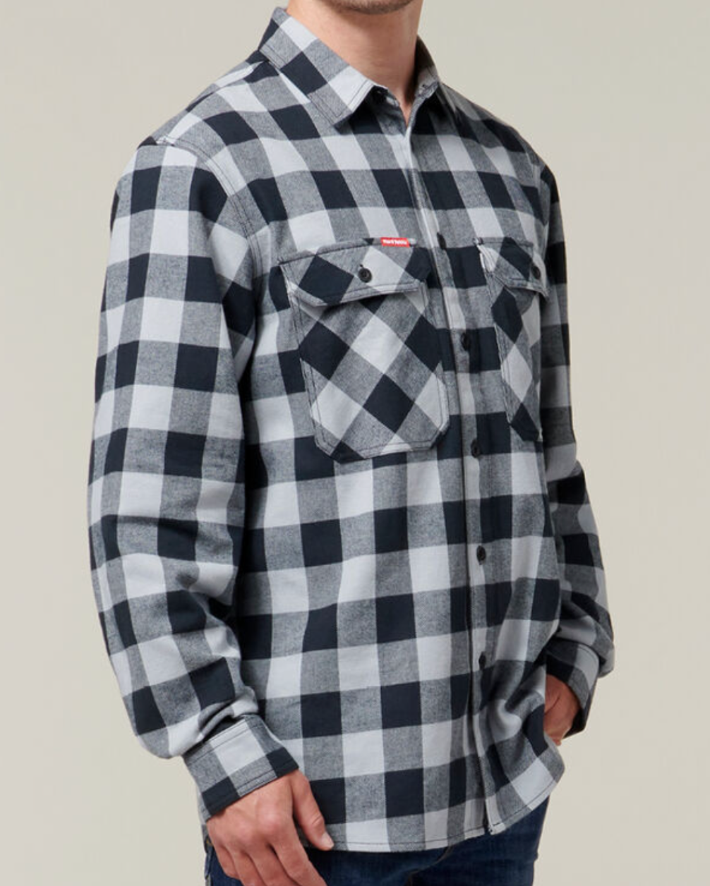 A person is seen from the neck down wearing a long-sleeved Hard Yakka Check Flannelette Shirt, characterized by its black-and-white checkered pattern and made of 100% Cotton Twill. The shirt features buttoned pockets on both sides of the chest and is paired with dark jeans.