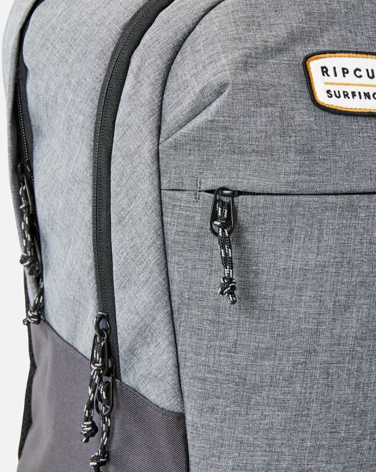 Close-up of the Rip Curl Overtime 30L Driven Backpack, showcasing a gray design with black zippers and a "Rip Curl Surfing Co." patch. Made from recycled polyester, it has a textured look and features black and white cord zipper pulls for a laptop-compatible layout.