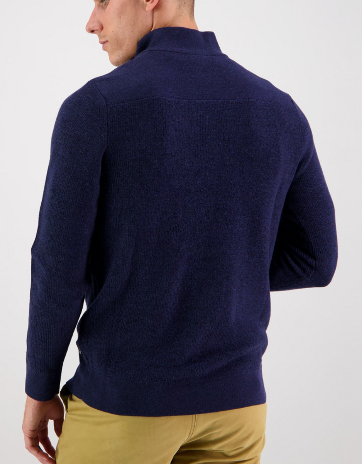 A man facing away from the camera wears a dark blue Swanndri Magnet Bay v2 Merino Jumper and tan pants against a plain white background.