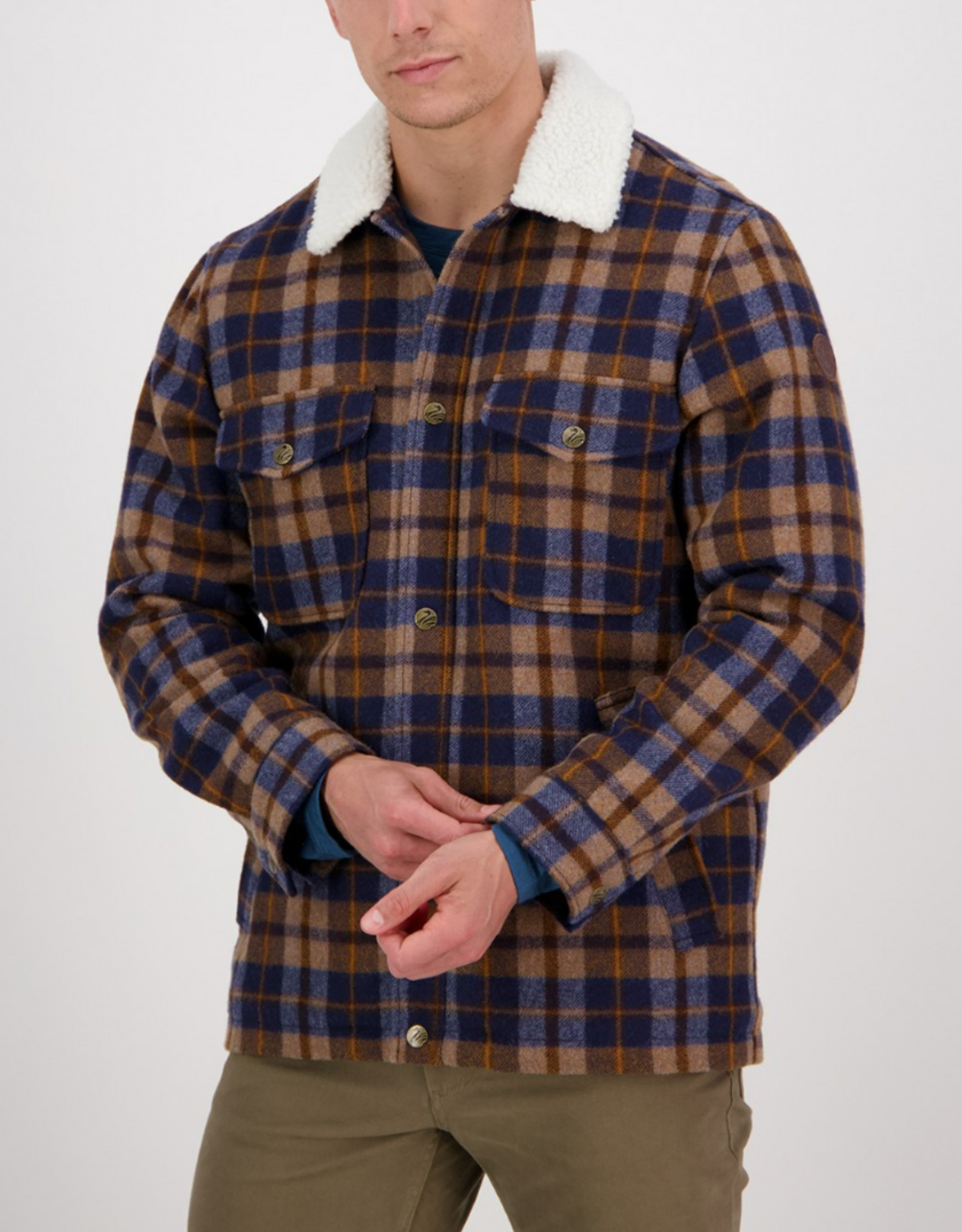 A person wears the Swanndri Kaituna Sherpa Lined Jacket in blue, brown, and beige plaid with a white sherpa collar, cozy sherpa lining, and buttoned chest pockets, standing against a plain background while adjusting their sleeve.