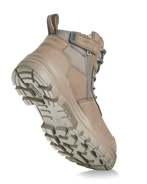 The Blundstone RotoFlex 8553 Mid Zip Safety Boot in beige is displayed from an angled bottom view. This boot showcases a side zipper, lace-up front, and a branded pull tab at the heel. The rugged, textured sole with a heavy-duty tread design is prominently visible, emphasizing its INFINERGY® cushioning technology.