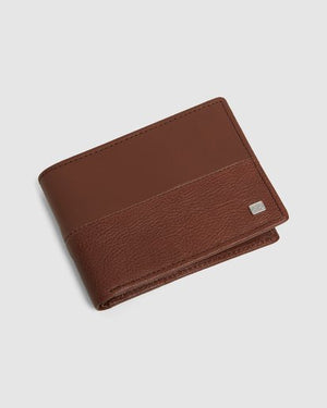 The Billabong Downtown RFID Slim-Line Wallet is pictured against a plain white background. The brown wallet features a smooth top section and a textured bottom section, separated by a horizontal seam. A small rectangular metal logo adorns the bottom right corner of the wallet.