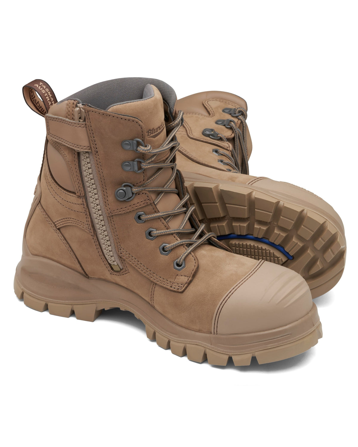 A pair of the Blundstone 984 Zip Sided Scuff Cap Safety Boots in tan, featuring rugged design with slip-resistant, thick, textured soles and convenient side zippers. One boot stands upright, displaying its laces and side zipper, while the other is tilted forward to reveal the sole pattern. Both boots come with reinforced steel toe caps and boast sturdy construction.
