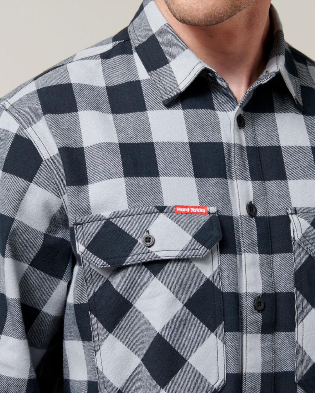 A close-up view of a person wearing a Hard Yakka Check Flannelette Shirt made from 100% Cotton Twill, featuring a black and white checkered pattern. The shirt includes two chest pockets, one adorned with a small red tag with white text. The individual is posed against a neutral background.