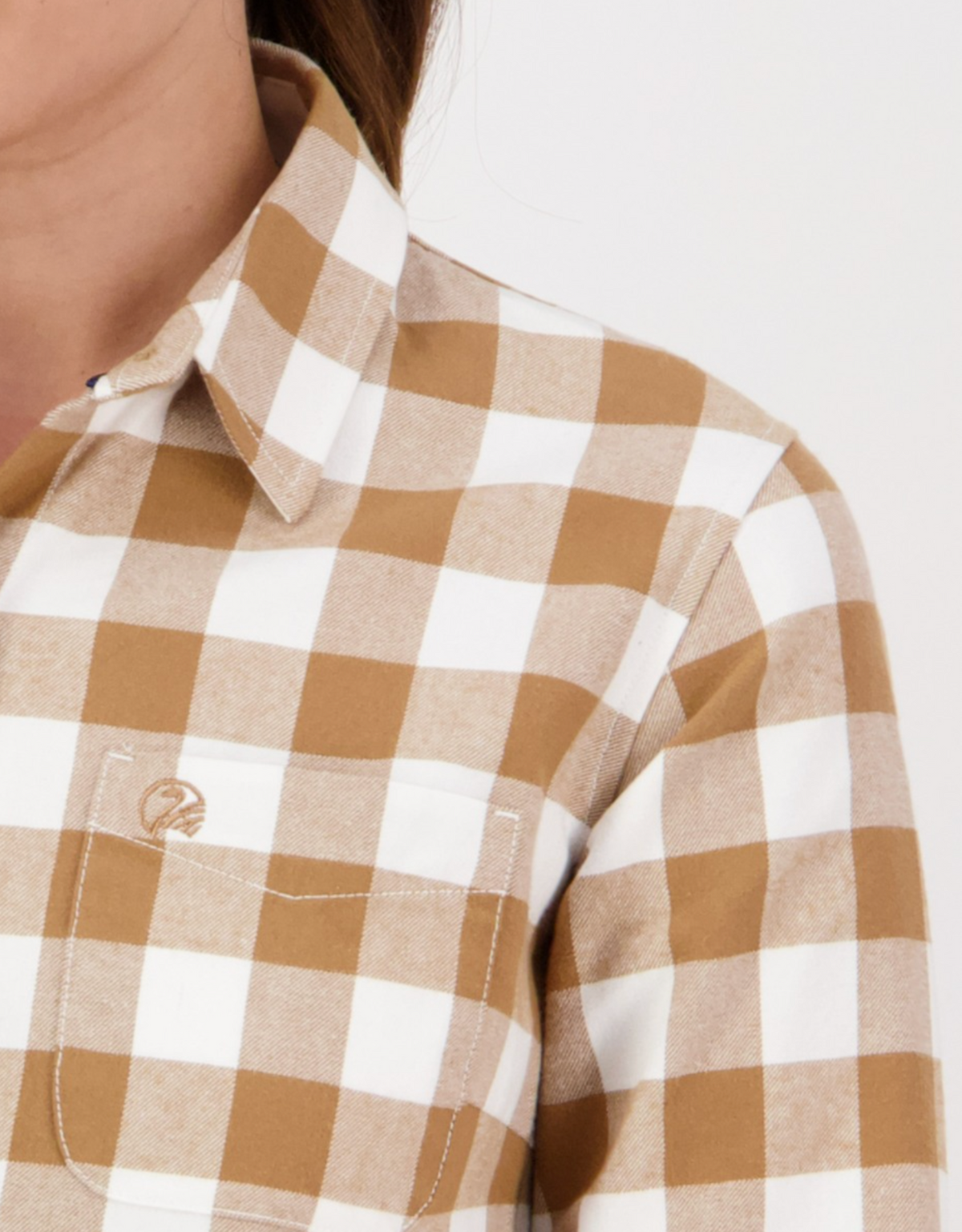 Close-up of a person wearing the Swanndri Womens Taranaki Tailor Shirt in tan and white check flannel, featuring a tailored fit, collar, front pocket with embroidered logo, and crafted from 100% cotton. Part of neck and shoulder visible.