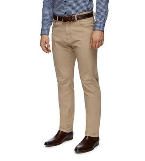 A person wearing a blue patterned shirt, beige City Club Austin Road Pant, and dark brown leather shoes stands casually with hands at their sides. The image focuses on the lower half of the body, showcasing the stylish pants and shoes.