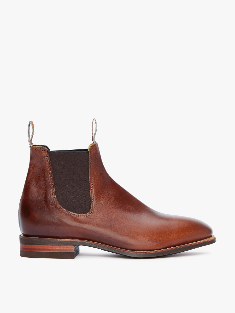 The RM Williams Comfort Craftsman Boot is a brown yearling leather Chelsea boot with a rounded toe, elastic side panels, front and back pull tabs, and a low stacked heel. Shown in side profile against a white background, inspired by the classic original design.