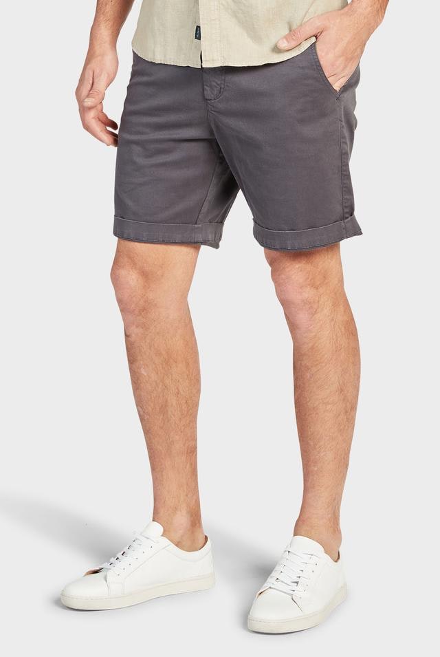 A person wearing a beige shirt paired with dark gray Academy Brand Cooper Chino Shorts and white sneakers. The person has their hands in their pockets and is standing with one leg slightly in front of the other against a plain white background.