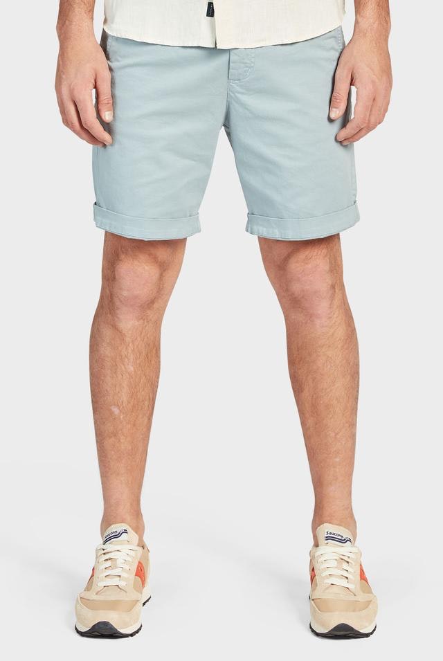 A person stands against a plain white background, wearing Academy Brand Cooper Chino Shorts in light blue cotton that are rolled up at the hem and a light-colored shirt. The person is also sporting beige and white sneakers with hints of orange. Only the person's legs and lower torso are visible.