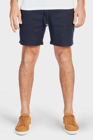 A person sports the Academy Brand Riviera Linen Short in navy blue with an elastic waist and relaxed fit, paired with camel sneakers. Their hands rest at their sides as they stand on a white background, wearing a white shirt; only the lower body is visible.