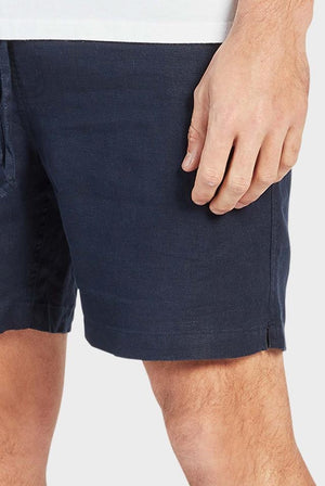A person stands against a plain background wearing Academy Brand Riviera Linen Shorts in dark blue. These elastic-waist shorts are relaxed for comfort, with their right hand casually by their side, showcasing visible skin. The simple design and lightweight fabric complete the look.