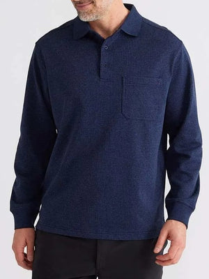 A man wears the Breakaway Micro Check Micro Prima Polo, a dark blue long-sleeve shirt with buttons, a chest pocket, textured fabric, and a relaxed fit. He pairs it with black pants against a plain light gray background.