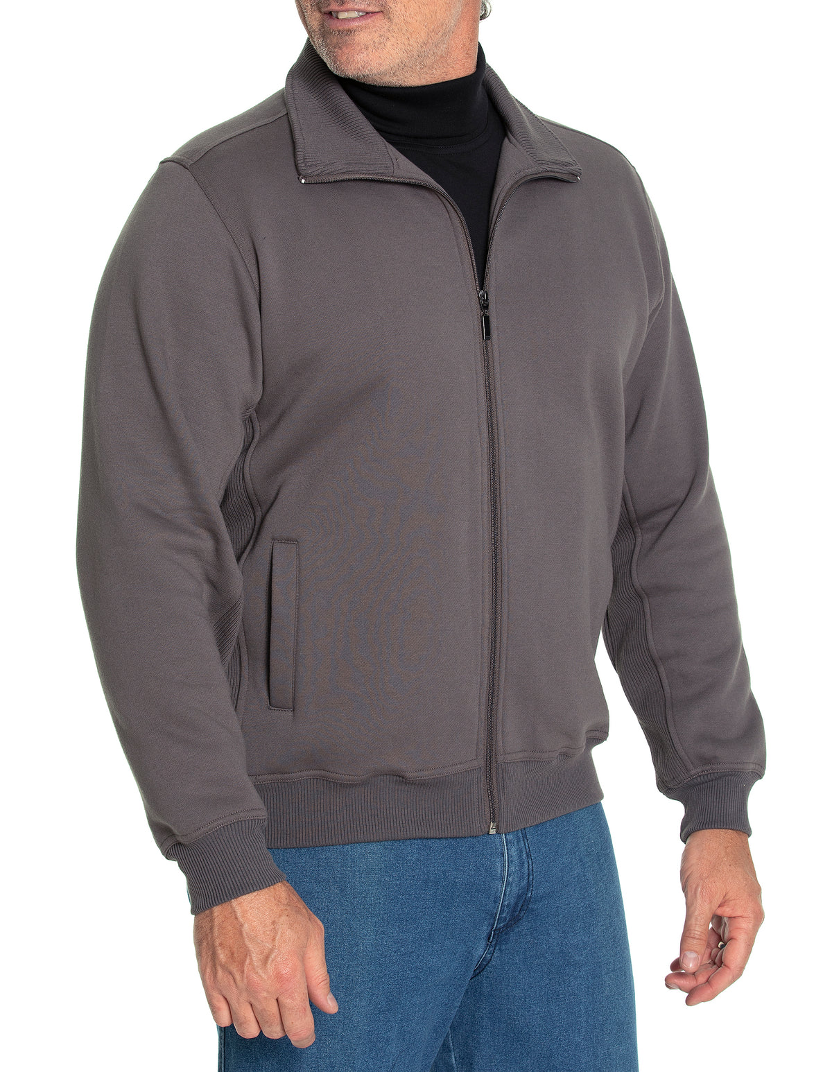 Wearing a "Breakaway Zed Snowy Mt Fleece Full Zip" in dark gray, someone stands against a white background with one hand in their pocket. The fleece jacket has rib knit cuffs and a high collar, while their face is partially obscured. They're also dressed in blue jeans.