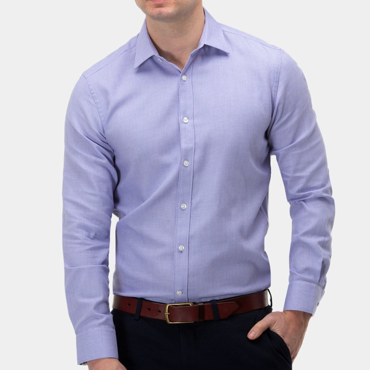 A person wearing a light purple Brooksfield Micro Textured Shirt, dark pants, and a brown belt with a gold buckle. The individual has one hand in a pocket, and the background is plain and light-colored.