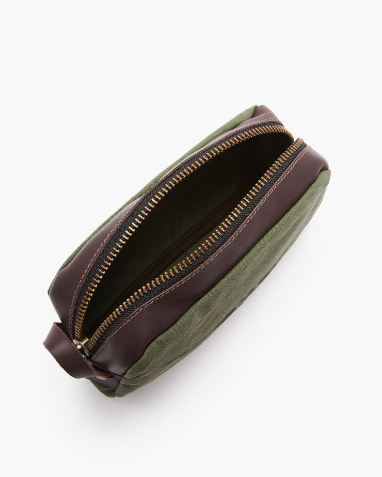 A top-down view of the RM Williams Ute Washbag with a green fabric body, dark brown leather trims, brass zipper, and a small leather handle at one end. The empty washbag sits on a white background.