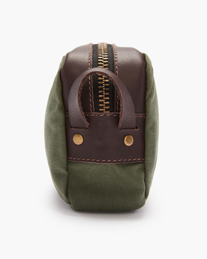 The RM Williams Ute Washbag is a small green canvas bag with brown leather trims, top zipper, brass rivets, stitching detail, and a side leather handle—showcasing classic RM Williams craftsmanship.