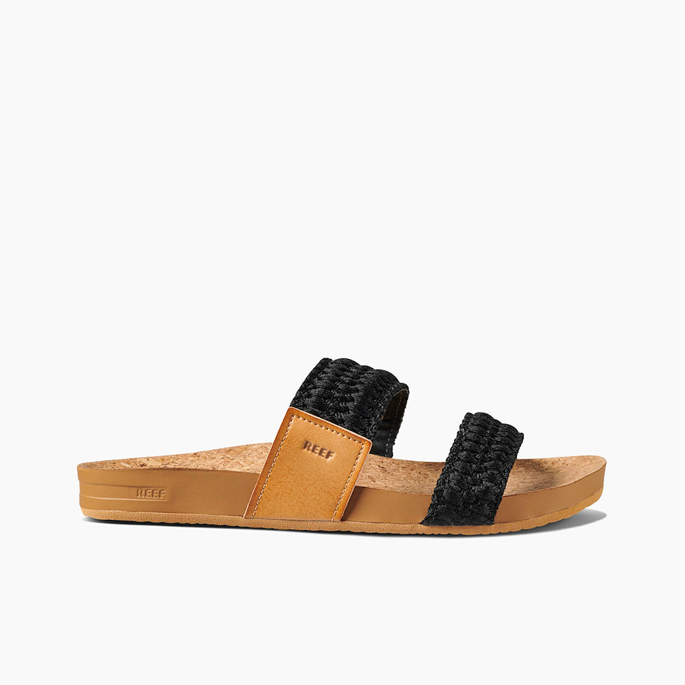 A side view of the REEF Cushion Vista Thread sandal, showcasing its tan cork and leather footbed with two black woven straps. The sandal features a cushion bounce midsole and has a flat sole with the "REEF" label on the side. The background is white.