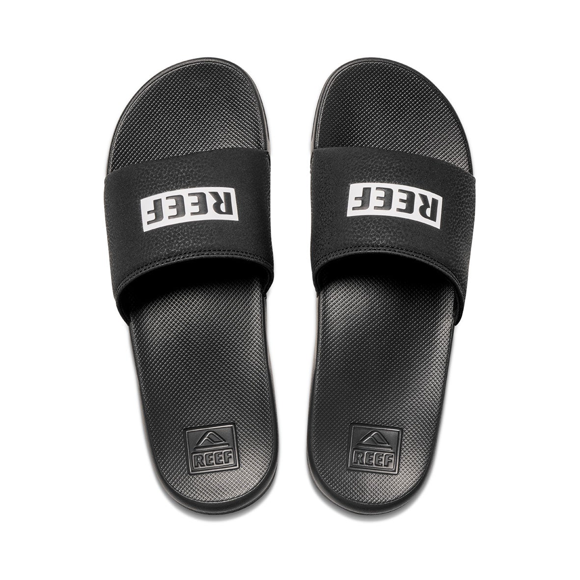 The REEF One Slide sandals are black with "REEF" in white on the straps, featuring Eco-One biodegradability. The textured soles have an embossed logo near the heel. These eco-friendly slides are positioned side by side, facing forward.