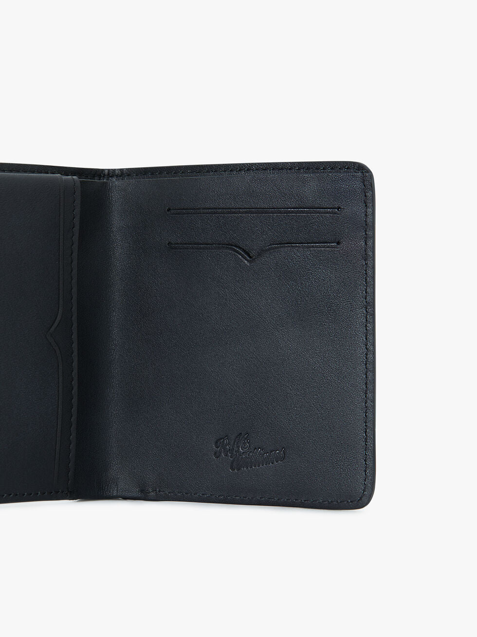 An RM Williams Urban Bi-Fold wallet in black, open to display its full-grain leather interior. The left side features a pocket, while the right side has two card slots. The wallet has embossed writing near the bottom right corner. The background is plain white.