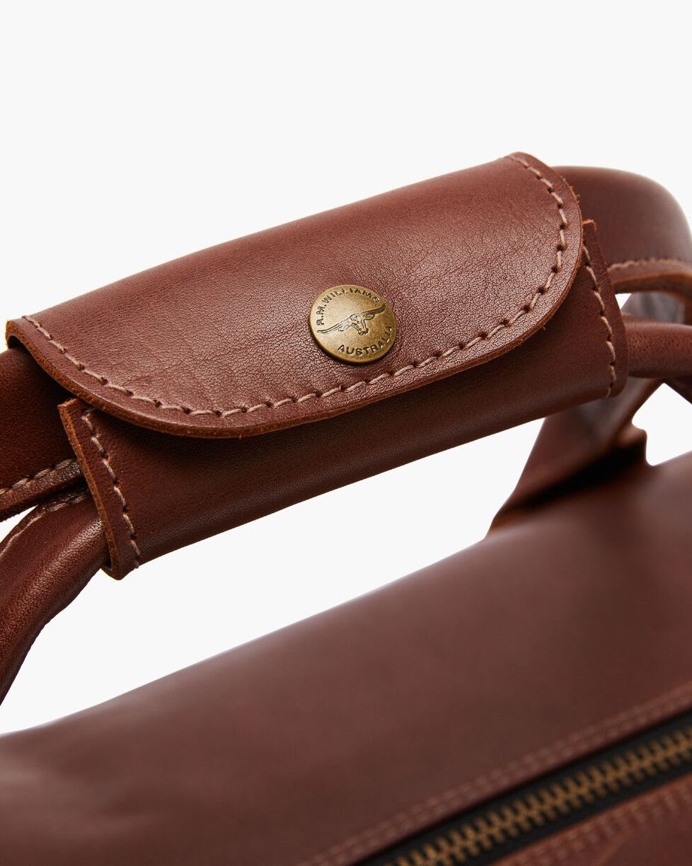 Close-up of the RM Williams Leather Ute Bag's brown leather handle, featuring a stitched flap and round brass "Australia" button. Visible zipper showcases its premium Australian Made craftsmanship.