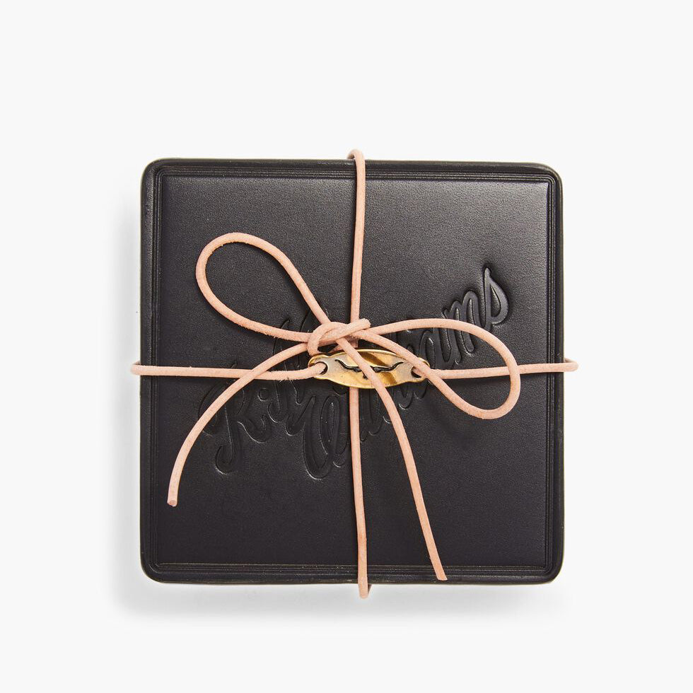 The RM Williams Heritage 6 Pack Coaster Set features a black square leather case with embossed text on the lid, tied with a beige leather cord in a bow, showcasing fine Australian craftsmanship against a white background.