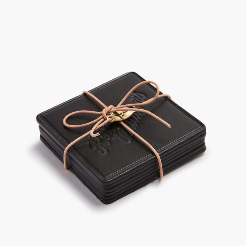 A black RM Williams Heritage 6 Pack Coaster Set is neatly tied with a brown leather cord and adorned with a small brass charm. These Australian-made coasters feature an embossed design, set against a plain white backdrop.