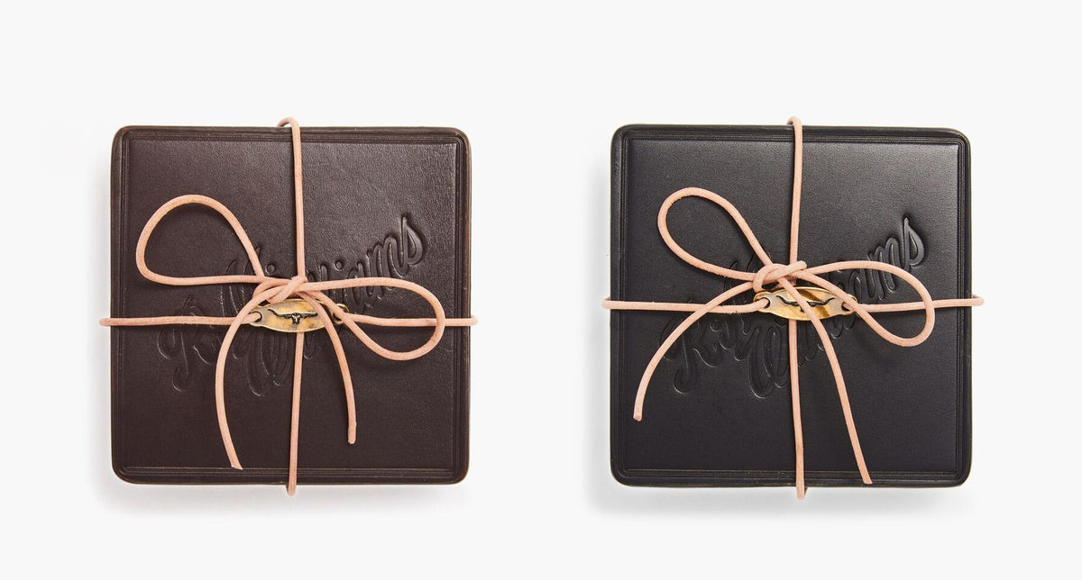 Two RM Williams Heritage coasters, one in dark brown and one in black leather, are embossed with the logo. Made in Australia, they are tied together with a light brown leather cord and placed side by side on a plain white background.